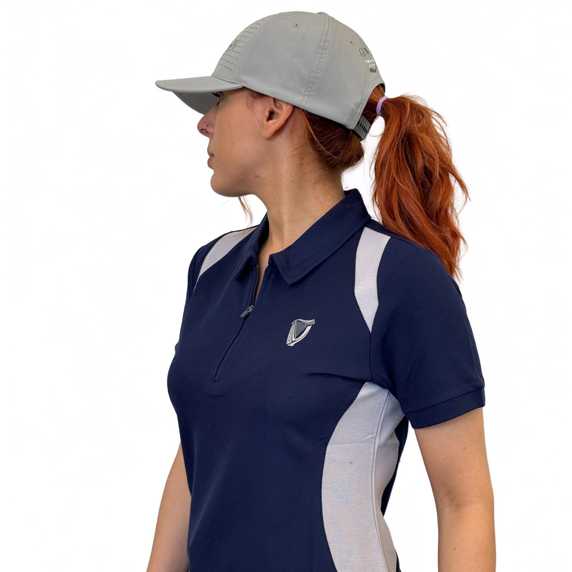 A woman wears the Ladies Performance Polo - Blue by Guinness Webstore US and a gray baseball cap, looking to the side against a plain white background.