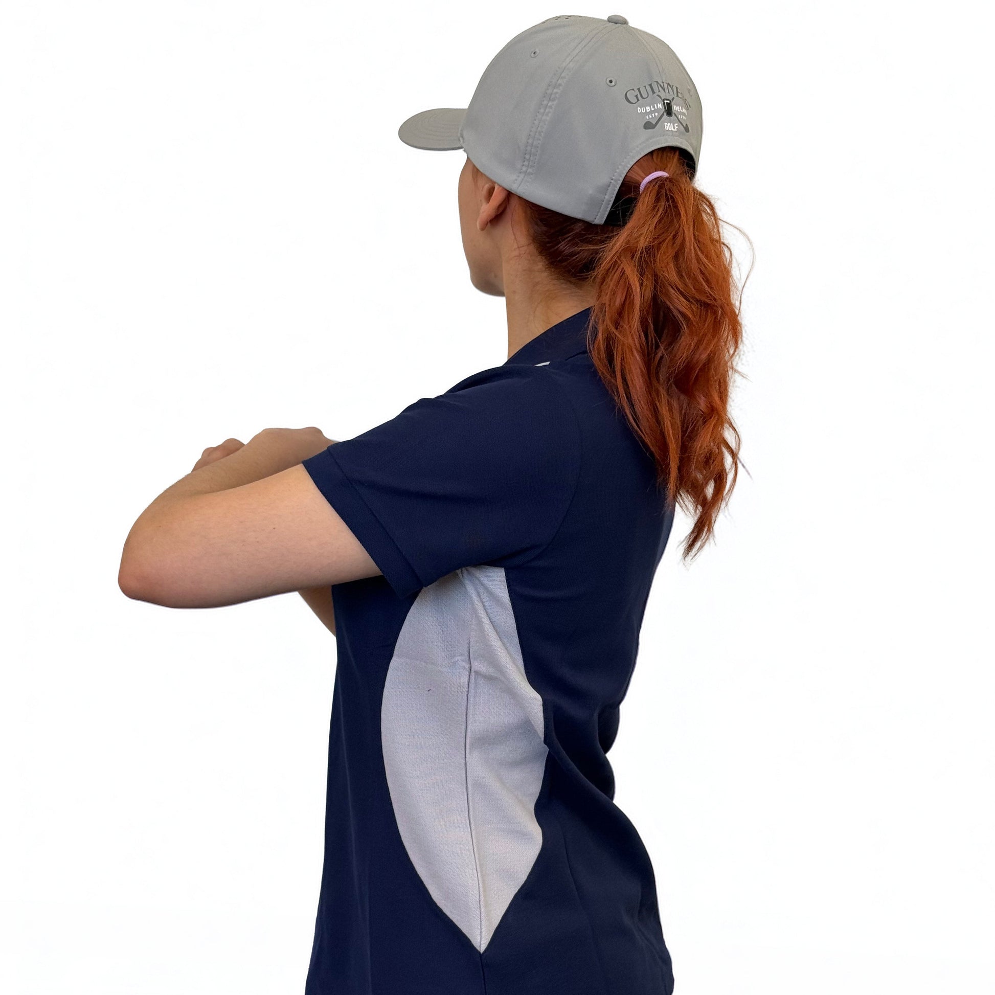 A woman with long red hair wears a gray cap, jeans, and the Ladies Performance Polo - Blue by Guinness Webstore US, standing sideways with arms crossed against a plain white background.