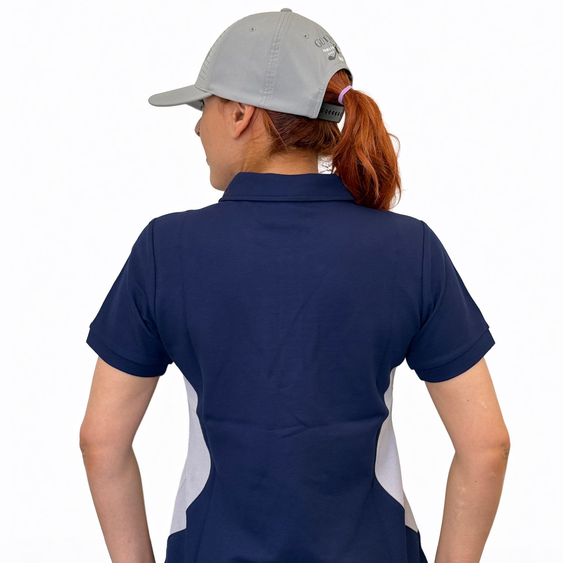 A person is shown from the back against a plain white background, wearing a grey baseball cap, light blue jeans, and the Ladies Performance Polo - Blue by Guinness Webstore US with white side panels.
