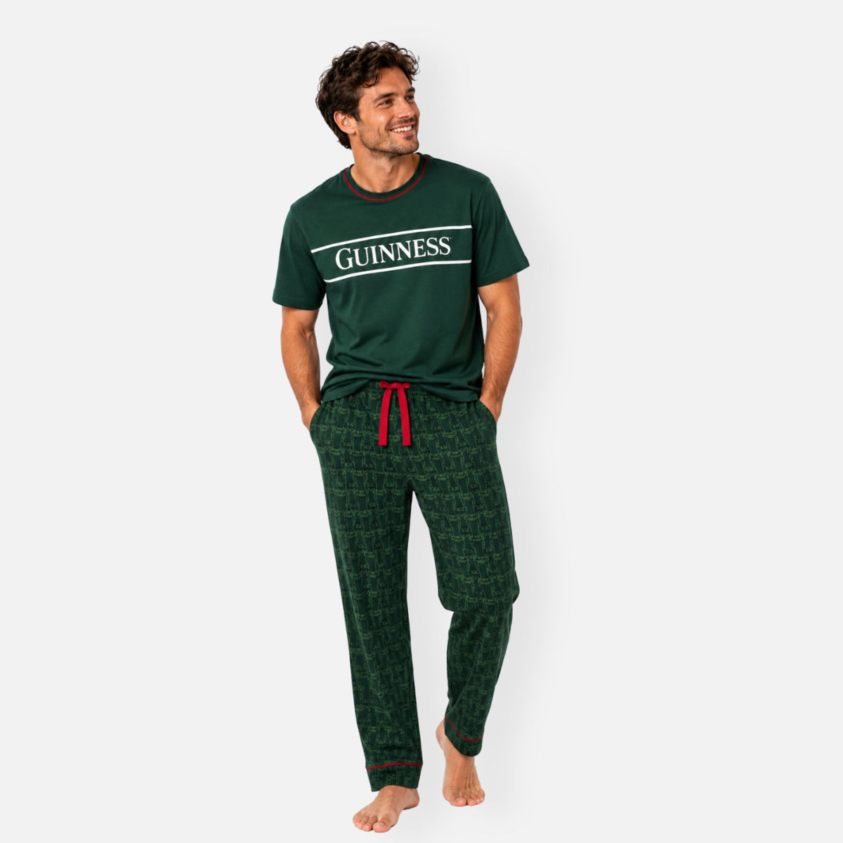 A man wearing a green Guinness Unisex 2-Piece Pajama Set featuring a T-shirt with “GUINNESS” on the chest and patterned lounge pants with a red drawstring.
