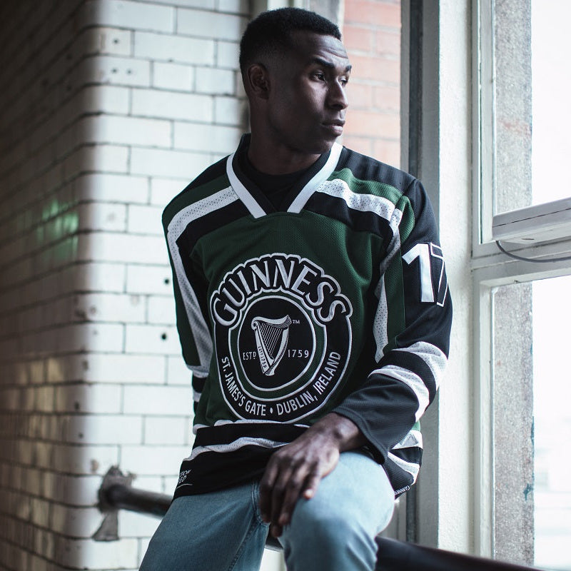 A man wearing a Guinness Green & White Hockey Jersey by Guinness sits by a window, looking outside.