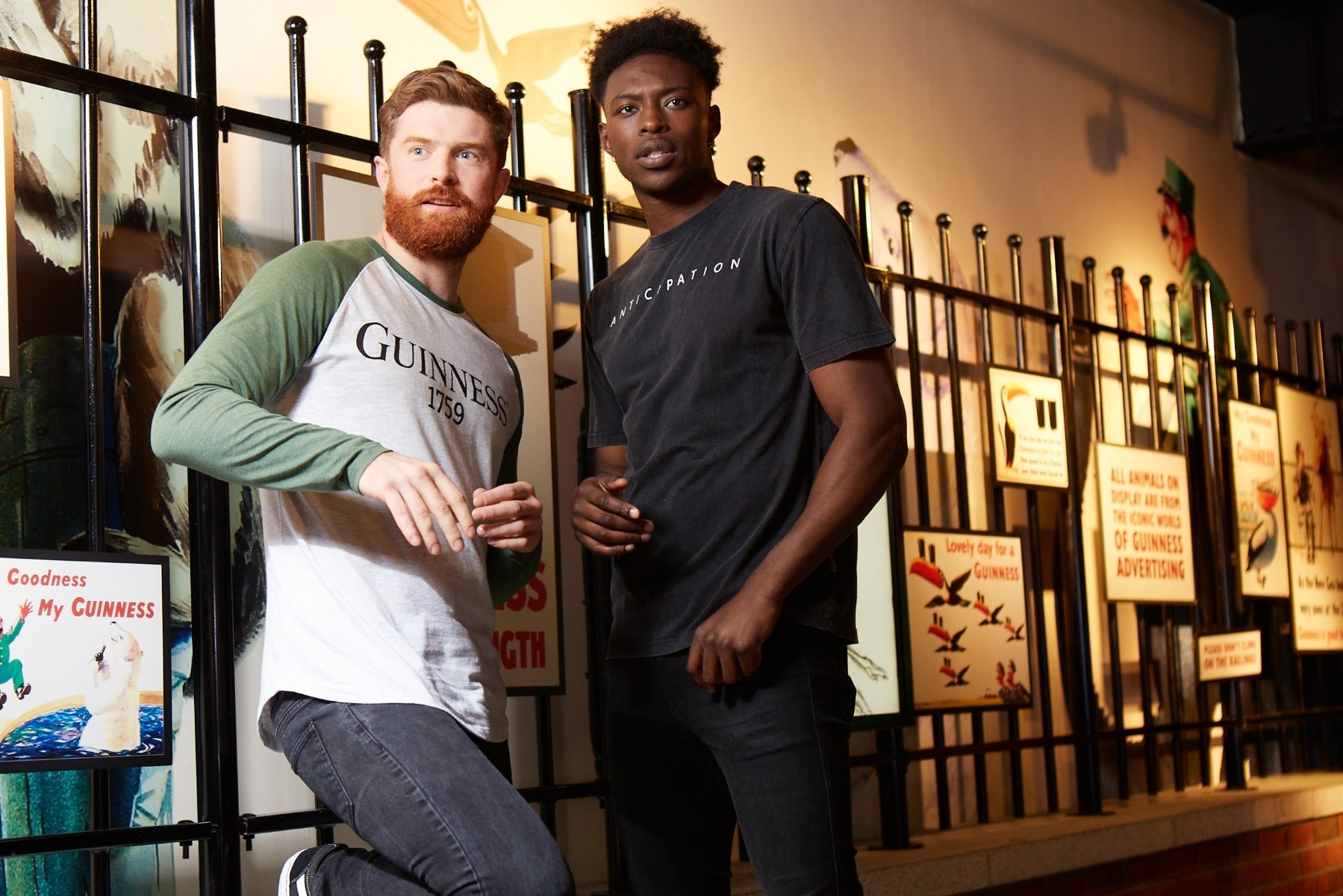 Two men stand indoors in front of a metal fence and a wall with vintage signs, one wearing the Guinness Premium Vintage Tortoise Back Graphic Tee featuring a subtle distressed look.