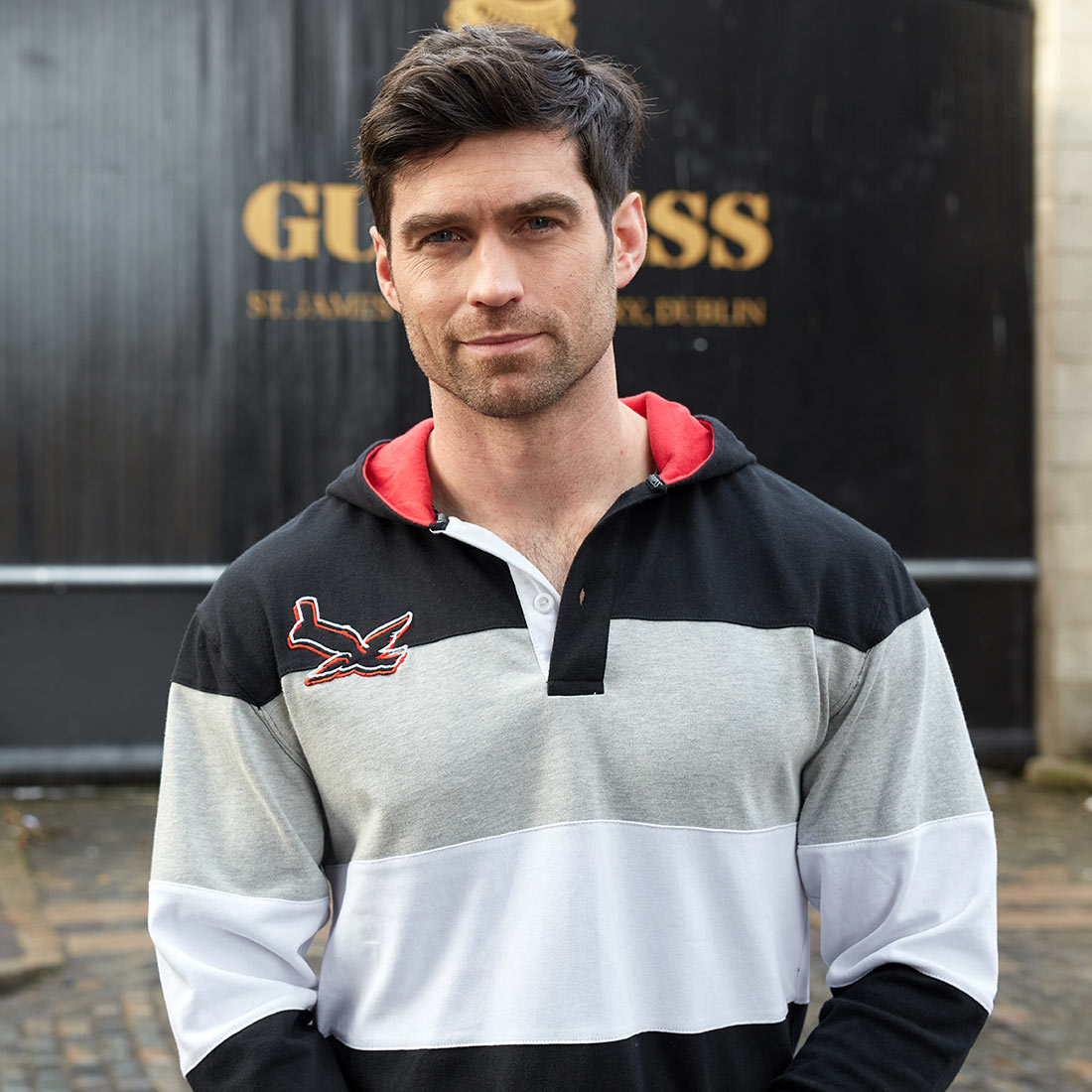 A man wearing a Guinness Black & Red Toucan Hooded Rugby jersey with grey, black, and red stripes.