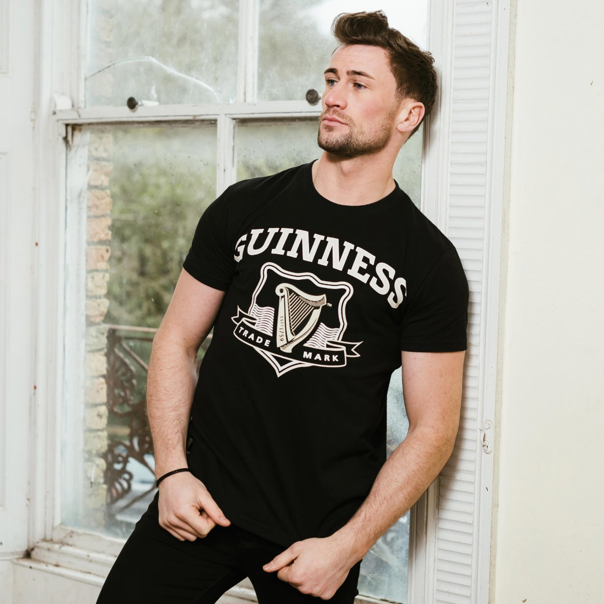 A man wearing a Guinness Signature Black Trademark Label Tee, featuring the Guinness® Trademark logo, leans against a window.
