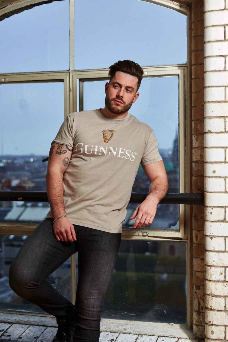 A man with a beard and tattoos leans against a windowsill, wearing a Beige Guinness Harp Premium Tee made of 100% cotton. He stands in front of a large window, with an urban landscape visible outside.