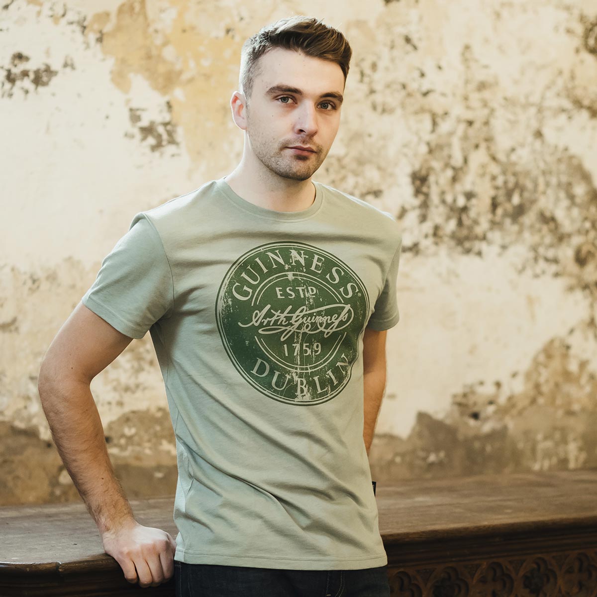 A man wearing a Guinness cotton blend green t-shirt leaning against a wall.