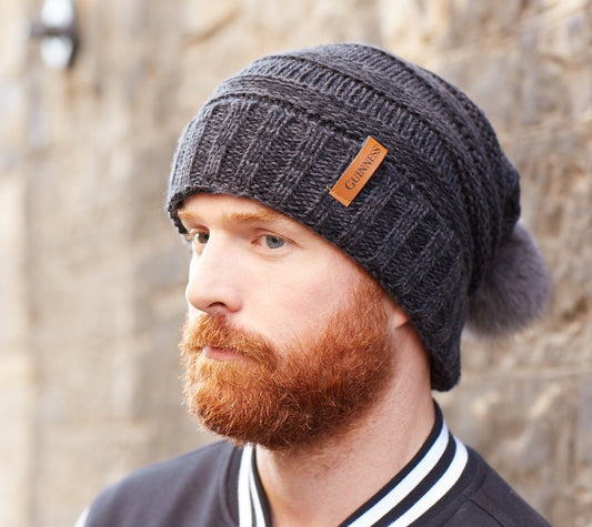 A bearded man wearing a beanie with a pom pom, styled in a Guinness Dark Grey Slouchy Bauble Beanie with Brown Leather Patch for the winter months.
