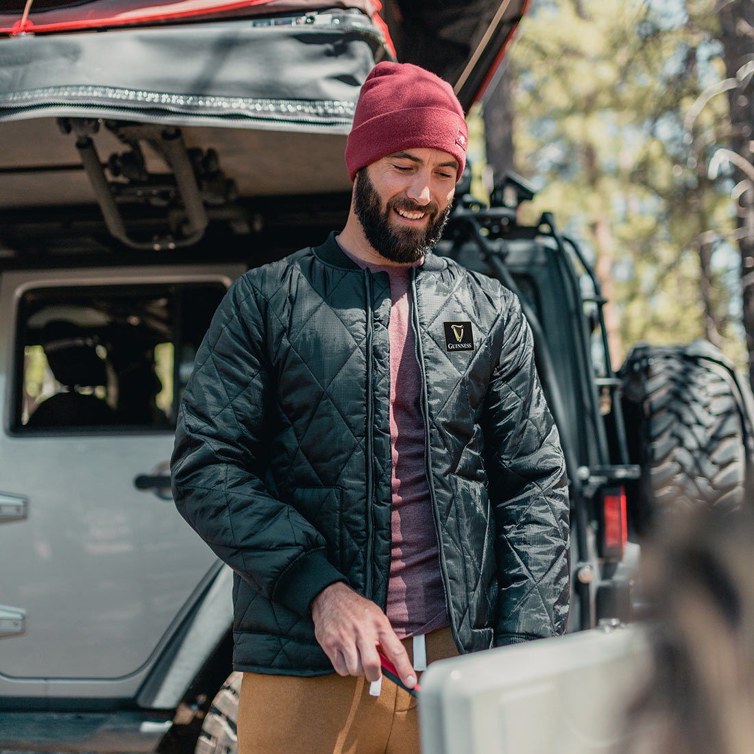 A man and woman wearing Guinness Quilted Utility Zip Up jackets standing next to a jeep in the woods.
