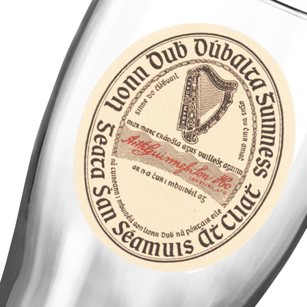 A close-up of the Guinness Vintage Label Pint Glass from Guinness Webstore US, showcasing the classic oval label with harp logo and Irish text. Available as a 2 Pack.