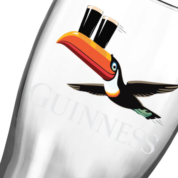 A close-up of the Guinness Gilroy Flying Toucan Pint Glass (24 Pack) from Guinness Webstore US, featuring "Guinness" and a flying toucan carrying two pints of beer on its beak.