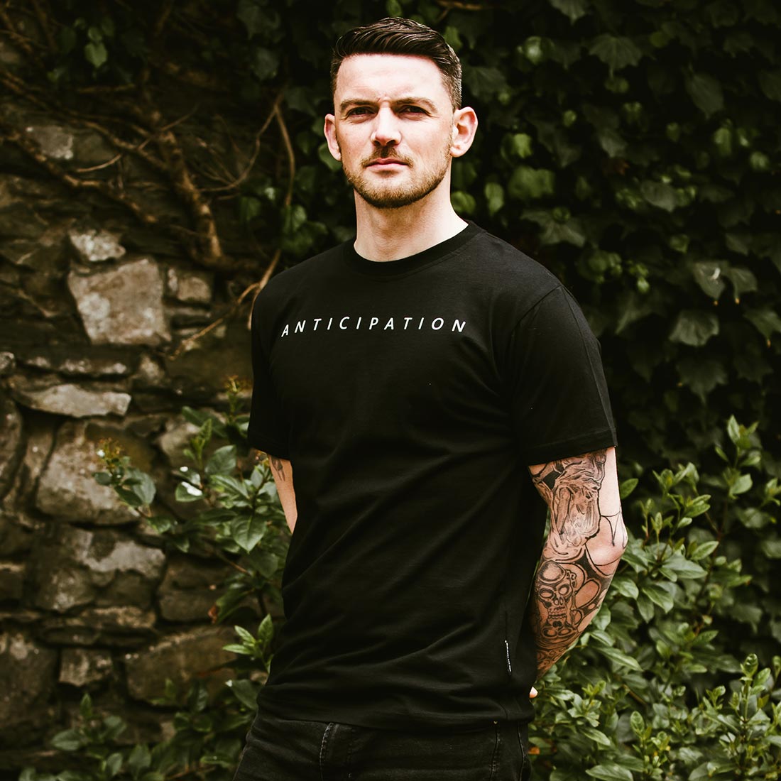 A man with tattooed arms stands outdoors before a stone wall and greenery, wearing the Guinness Premium Vintage Tortoise Back Graphic Tee, made from 100% cotton and featuring "ANTICIPATION" on the front.