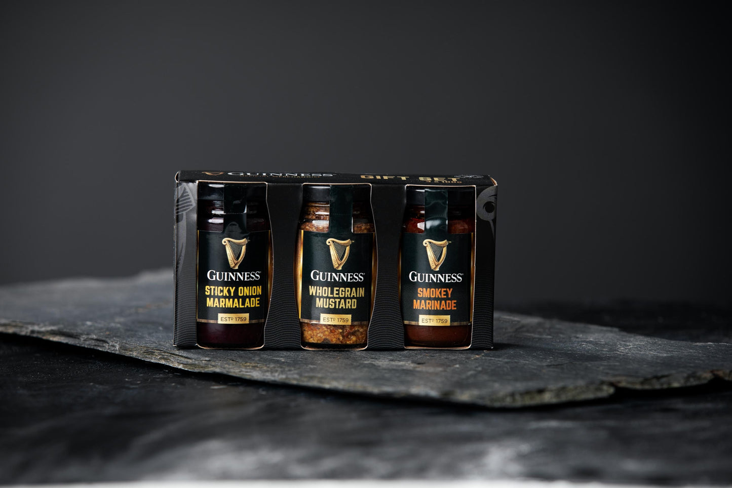 The Guinness Gourmet Gift Set 3 x 100g, featuring a trio of Guinness-branded condiments—Sticky Onion Marmalade, Wholegrain Mustard, and Smokey Marmalade—is elegantly showcased in black packaging on a dark slate surface. Perfect for any occasion, this gift set has been recognized by the Irish Food Awards.
