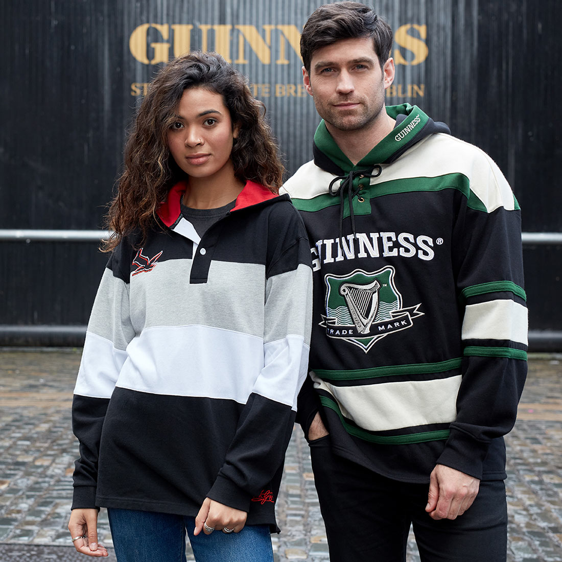 A man and a woman, wearing Guinness Black & Red Toucan Hooded Rugby jerseys, are standing in front of a Guinness sign.