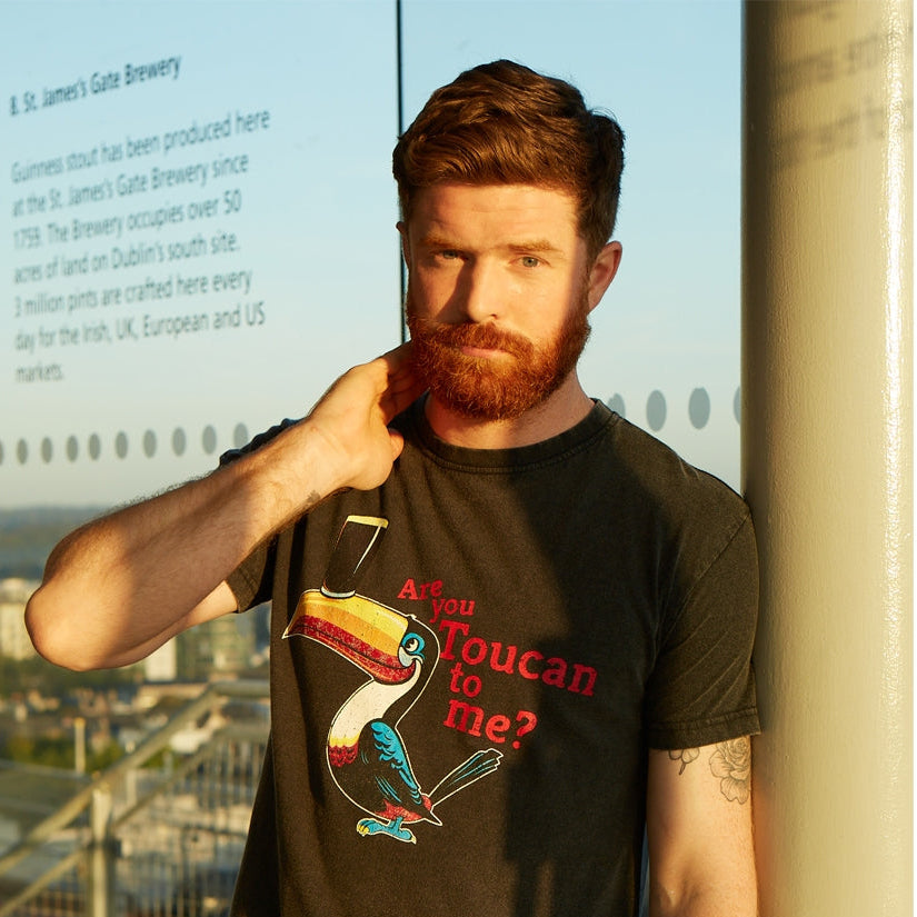 A man with a beard stands indoors near a window, wearing the Guinness Are You Toucan To Me Tee in grey, featuring a colorful toucan and playful text.