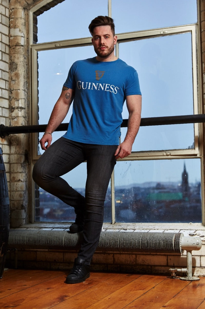 A man wearing a blue Guinness Trademark Label T-Shirt Blue in front of a window.