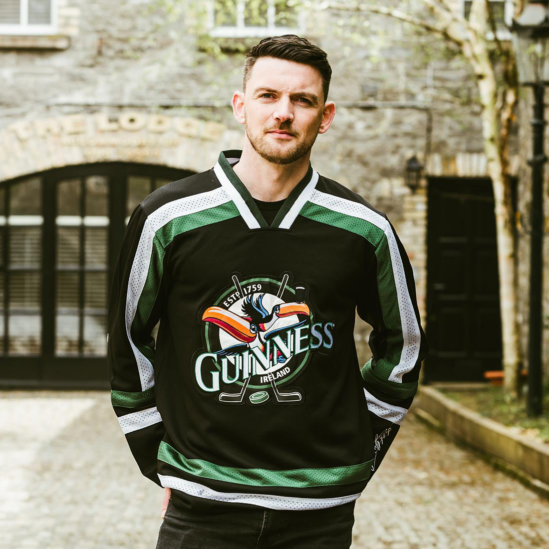 A man stands outside a stone building with his hands in his pockets, wearing the Guinness Toucan Hockey Jersey in black and green by Guinness.