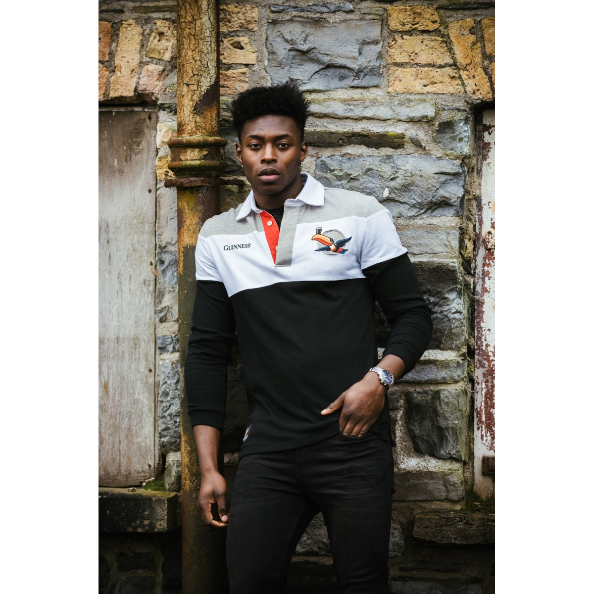 A young man leaning against a brick wall wearing a polo shirt, representing Guinness with the Guinness Toucan Rugby Jersey on his rugby jersey.