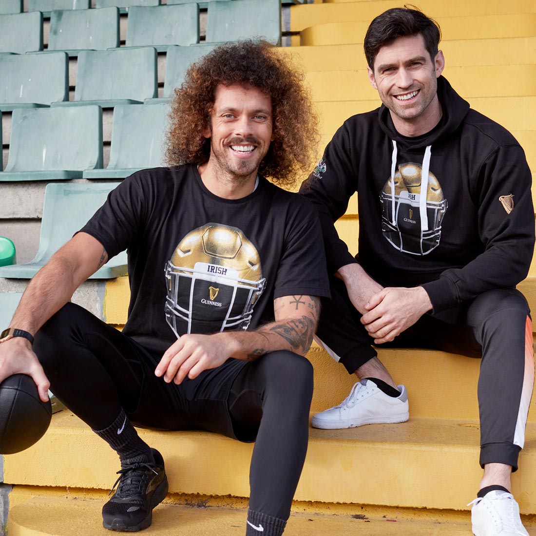 Two men sitting on the steps of a stadium, one wearing a Guinness Notre Dame Helmet T-Shirt Black and the other holding a Guinness.