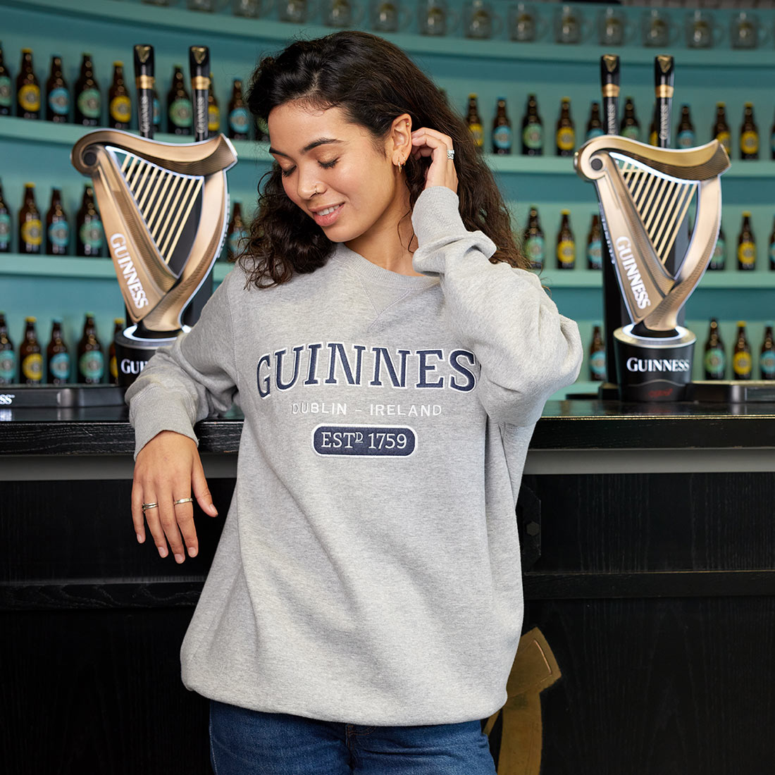 A woman wearing a Guinness Grey Crew Neck Sweatshirt leaning against a bar in Dublin, Ireland.