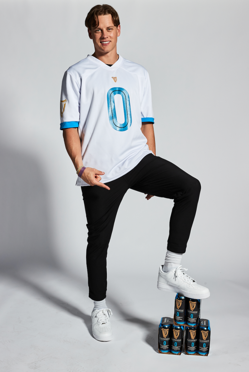 A white man posing next to a stack of Guinness 0 Football Jersey - White cans from the Guinness Webstore US.