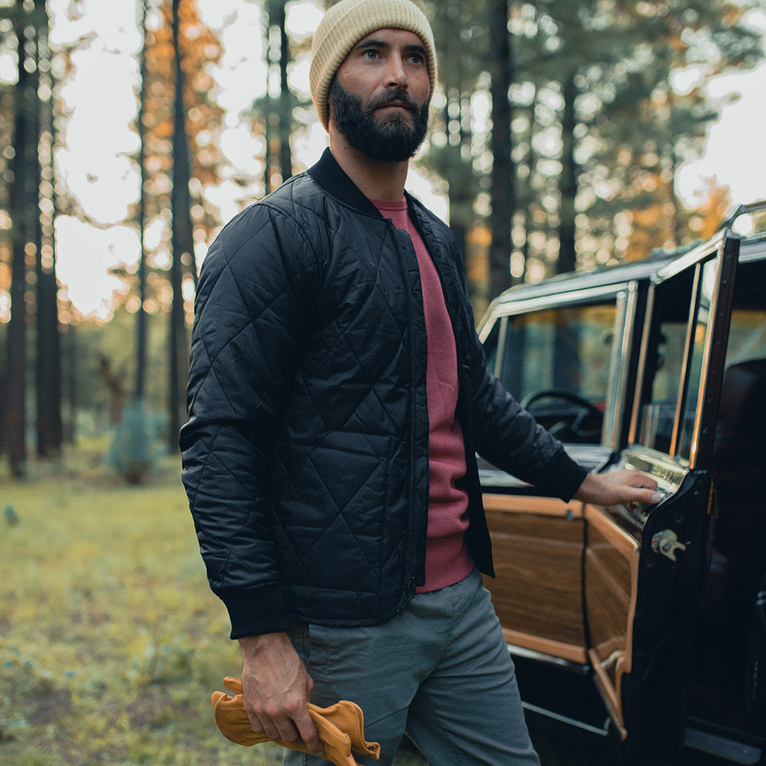 A waterproof Guinness bearded man standing next to a car in the woods, wearing a Guinness Quilted Utility Zip Up jacket.