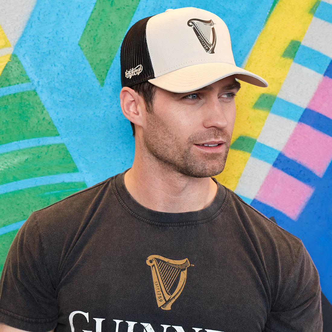 A man wearing a Guinness Premium Beige & Black Harp Hat in front of a colorful wall.