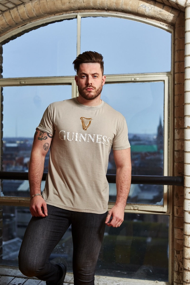A man with tattoos is standing indoors by a large window, wearing a Beige Guinness Harp Premium Tee made of 100% cotton and black jeans.