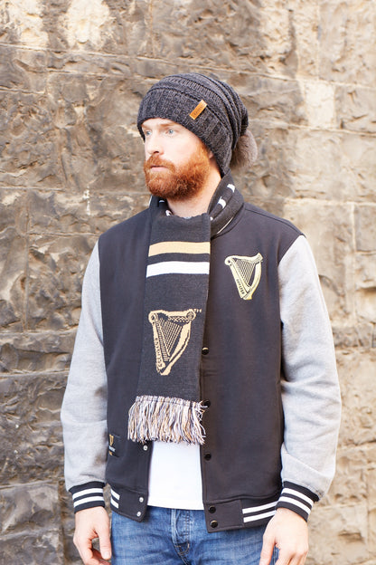 A bearded man wearing a Guinness Dark Grey Slouchy Bauble Beanie with Brown Leather Patch and 100% acrylic scarf.