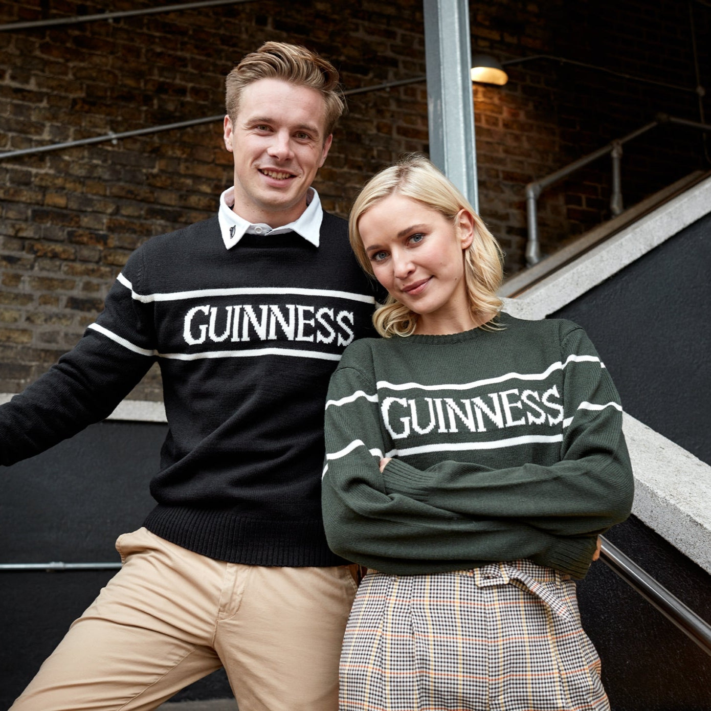 Two people wearing "Guinness Bottle Green Crew Neck Jumpers" from the Guinness Webstore US, featuring the iconic Harp logo, pose casually in front of a brick wall and stairway.