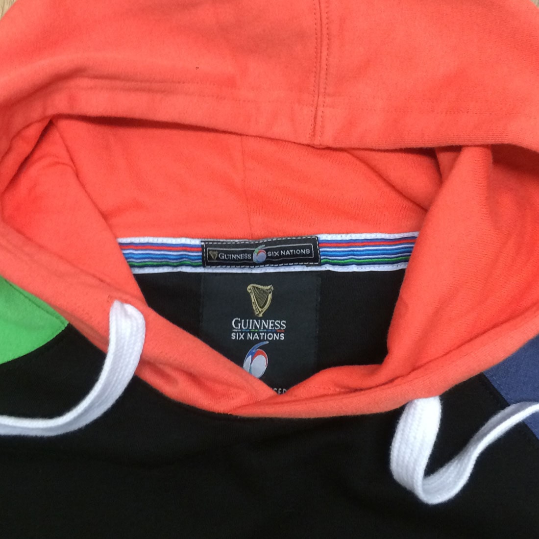Close-up of a clothing label with the text "Guinness® Six Nations" inside the collar of an orange and black Guinness Six Nations Color Block Hoodie.