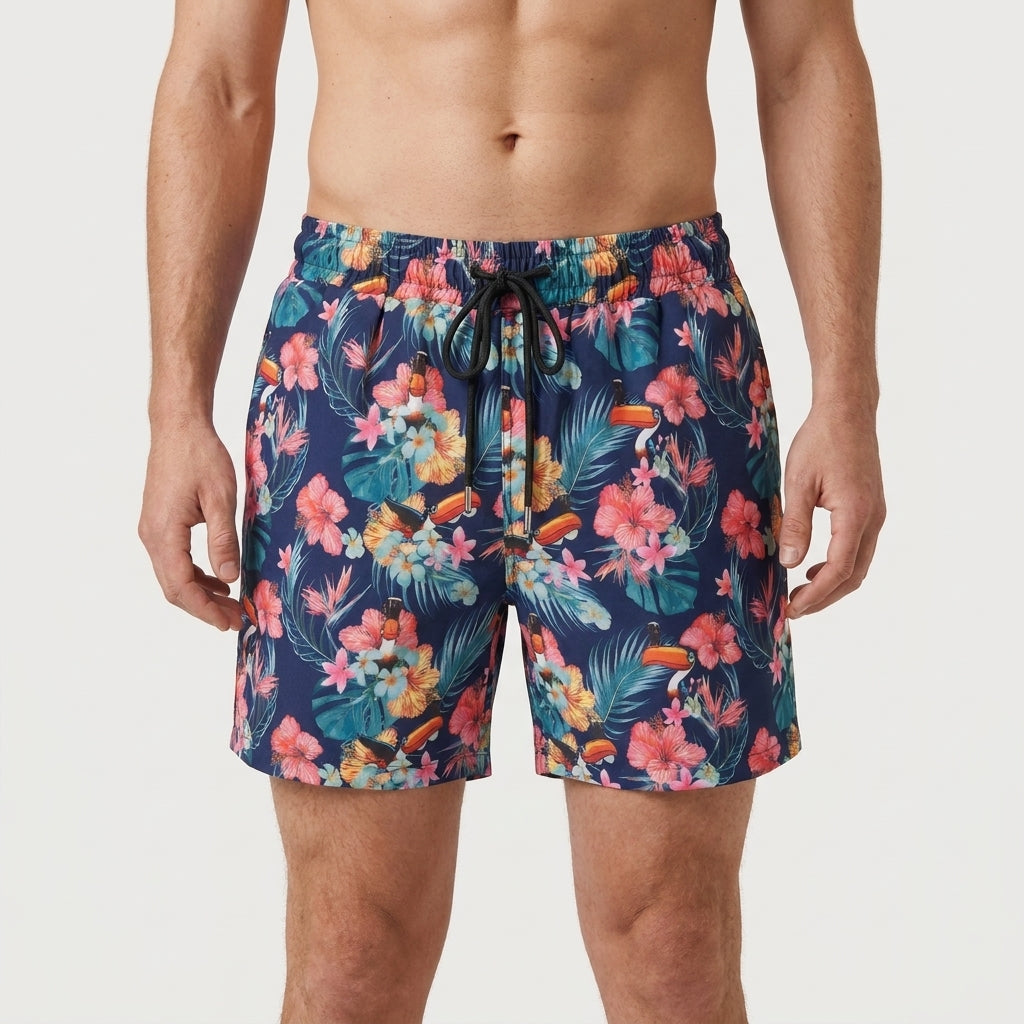 A man wears Guinness Toucan Hawaiian Beach Shorts by Guinness Webstore US—eco-friendly blue shorts with a vibrant tropical floral and leaf print and a drawstring waist, standing against a plain background.