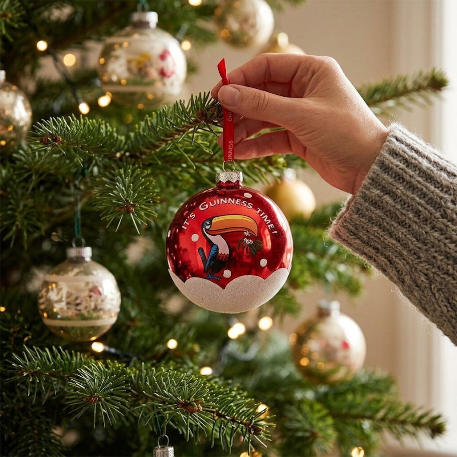 A hand hangs a Guinness Toucan Glass Bauble on a decorated Christmas tree, surrounded by other ornaments and twinkling lights in the background—a perfect festive addition from Guinness.