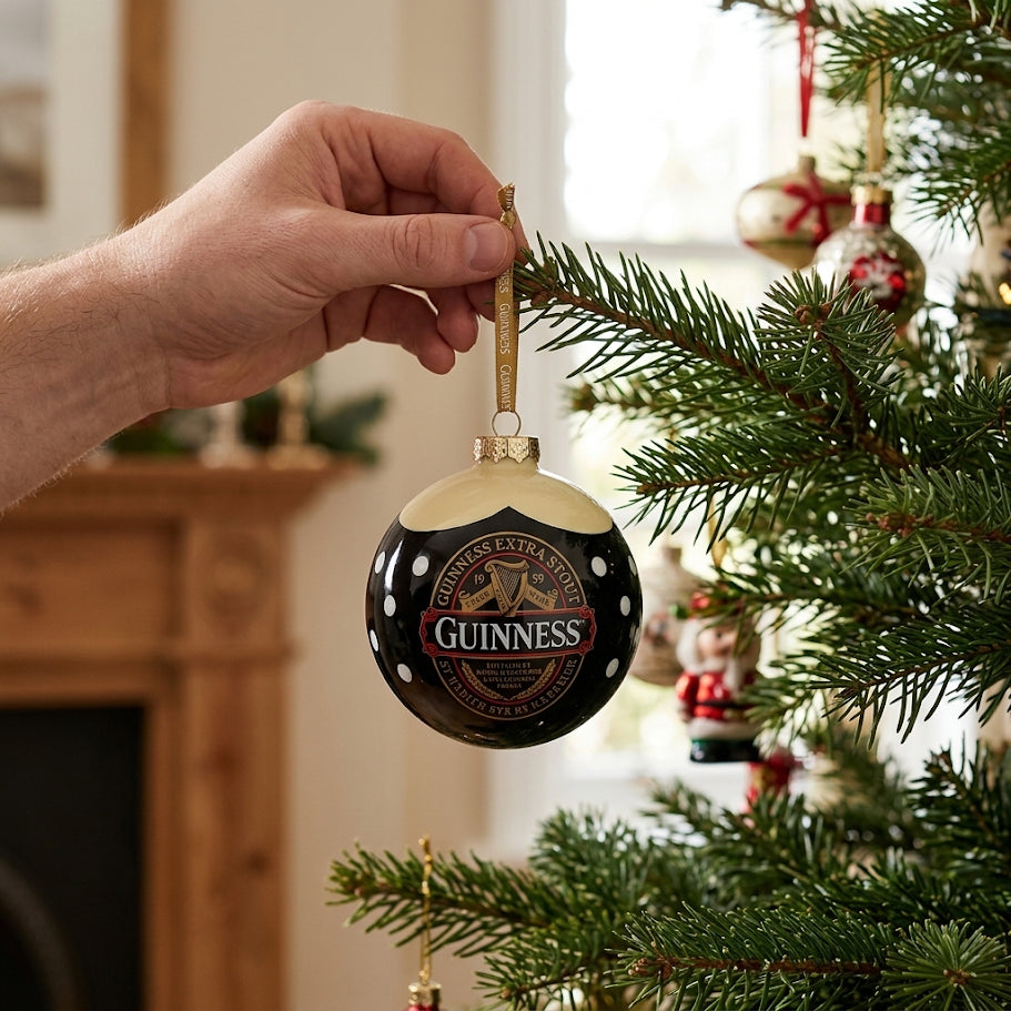 A hand hangs a Guinness Classic Label Glass Bauble on a decorated tree, with a fireplace and holiday decorations in the background—making it the ideal festive gift for any Guinness lover.