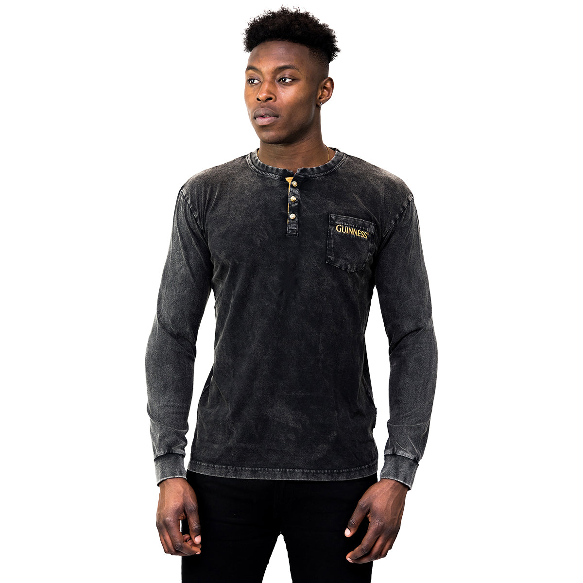 A man faces forward wearing the Guinness Classic Washed Black Henley by Guinness, featuring "Guinness" on the chest pocket. He pairs it with black pants against a plain white background.