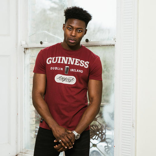 A man wearing a comfortable Guinness Signature Burgundy Trademark Label Tee in front of a window.