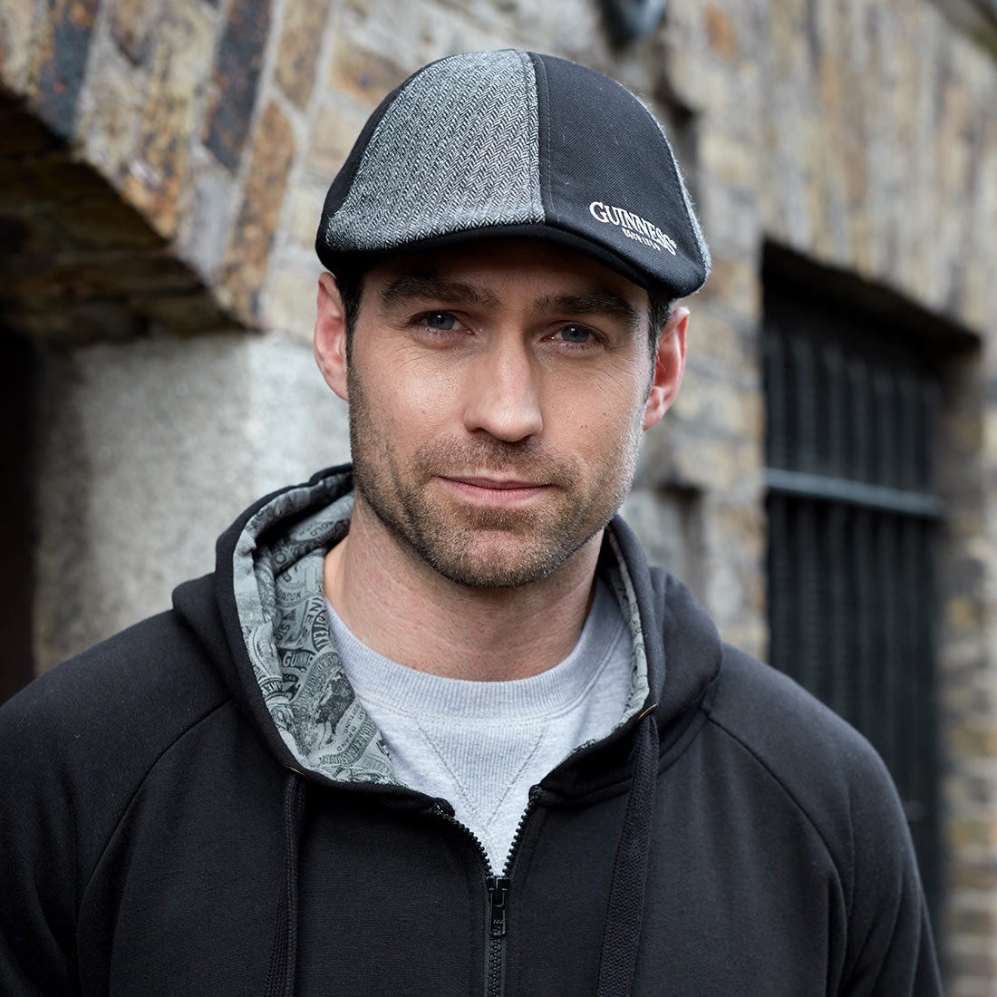 A man wearing a black hoodie, an embroidered Guinness paneled Ivy cap, and a hat.