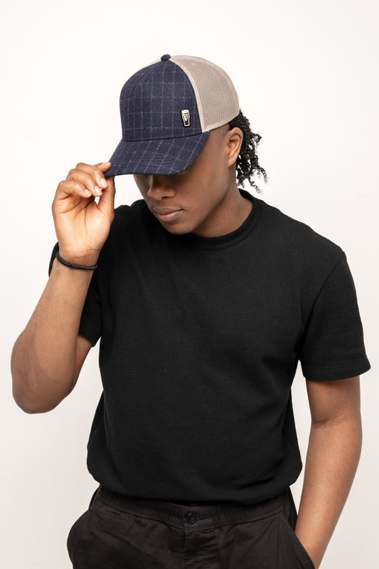 A person in a black t-shirt and dark pants adjusts a Guinness Checked Trucker Cap from Guinness Webstore US, looking down against a plain white background.