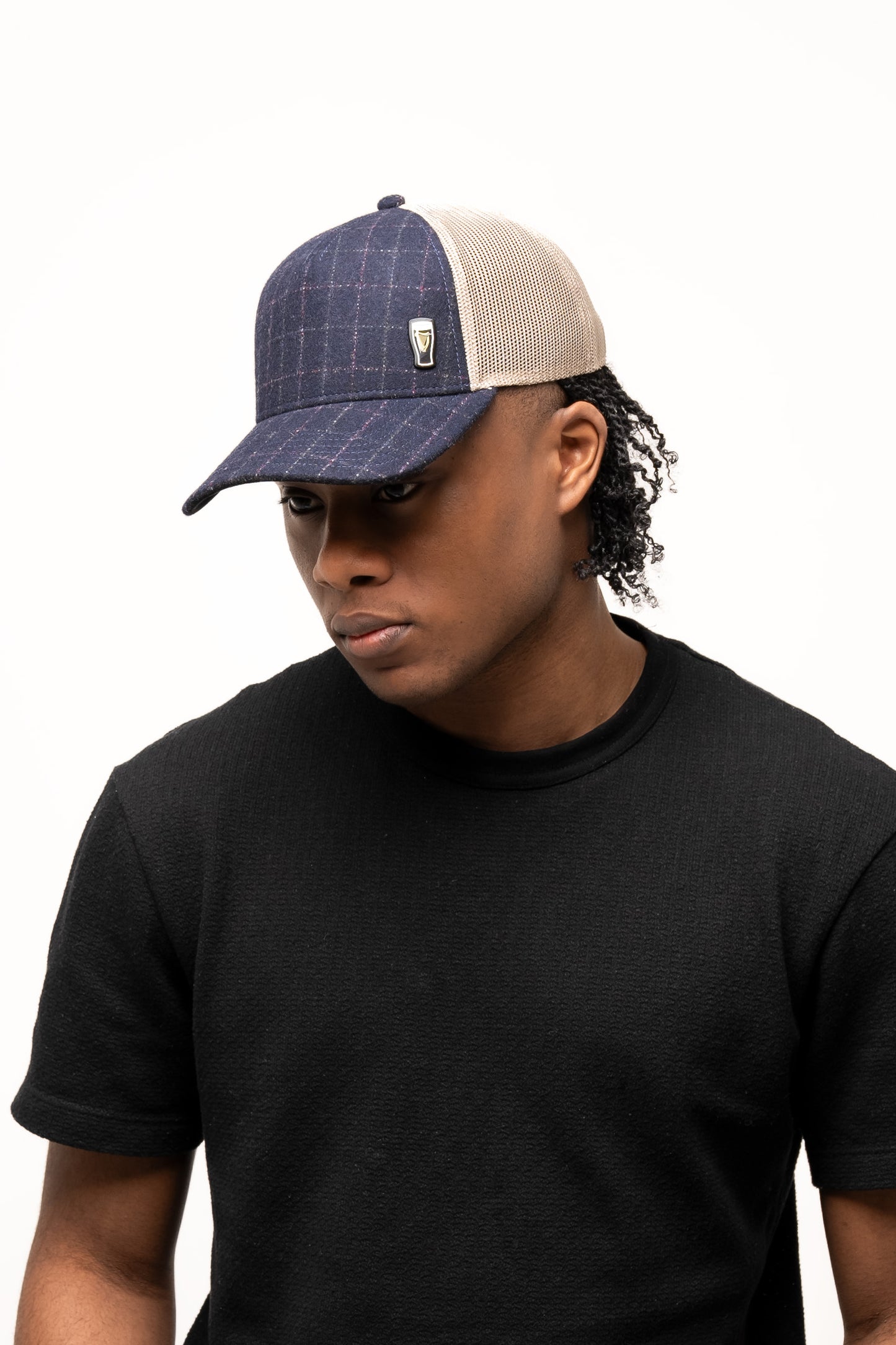 A person in a black t-shirt and a Guinness Checked Trucker Cap from Guinness Webstore US, with mesh back, faces slightly downward against a plain white background, subtly nodding to Irish heritage.