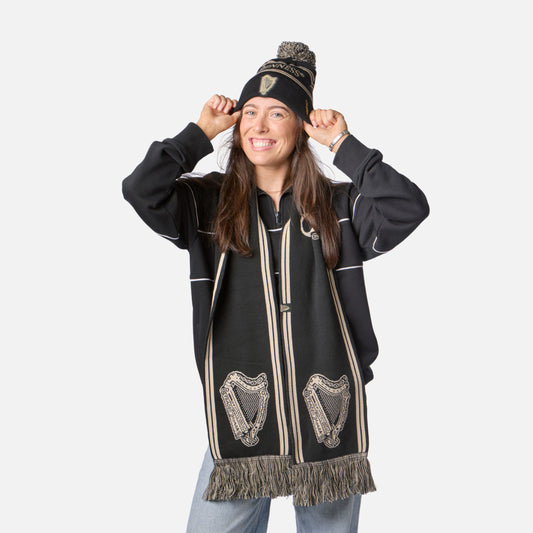 Woman smiling, wearing the Guinness Black & Gold Sports Beanie – Vintage Harp Knit, black scarf with harp designs, black sweater, and light blue jeans against a white background.