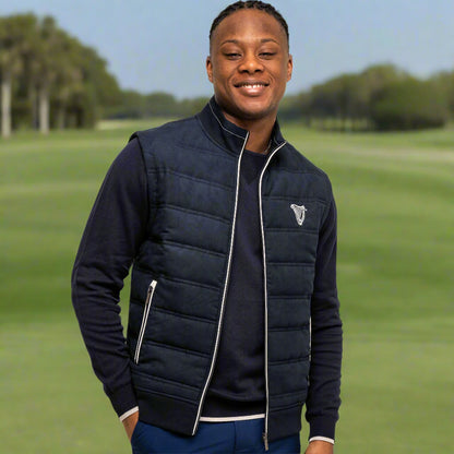 A man smiles in front of a white background, wearing a navy Guinness Performance Gilet from Guinness Webstore US over a dark long-sleeve shirt and blue shorts.