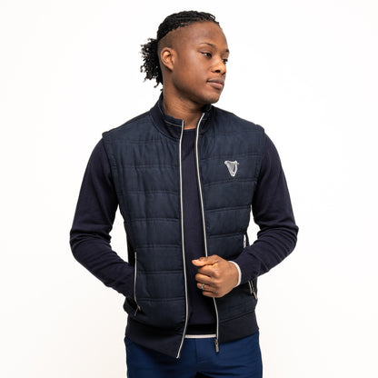 A person models the navy Guinness Performance Gilet by Guinness Webstore US, featuring quilted detailing, worn over a dark long-sleeve shirt and blue shorts, standing against a white background.