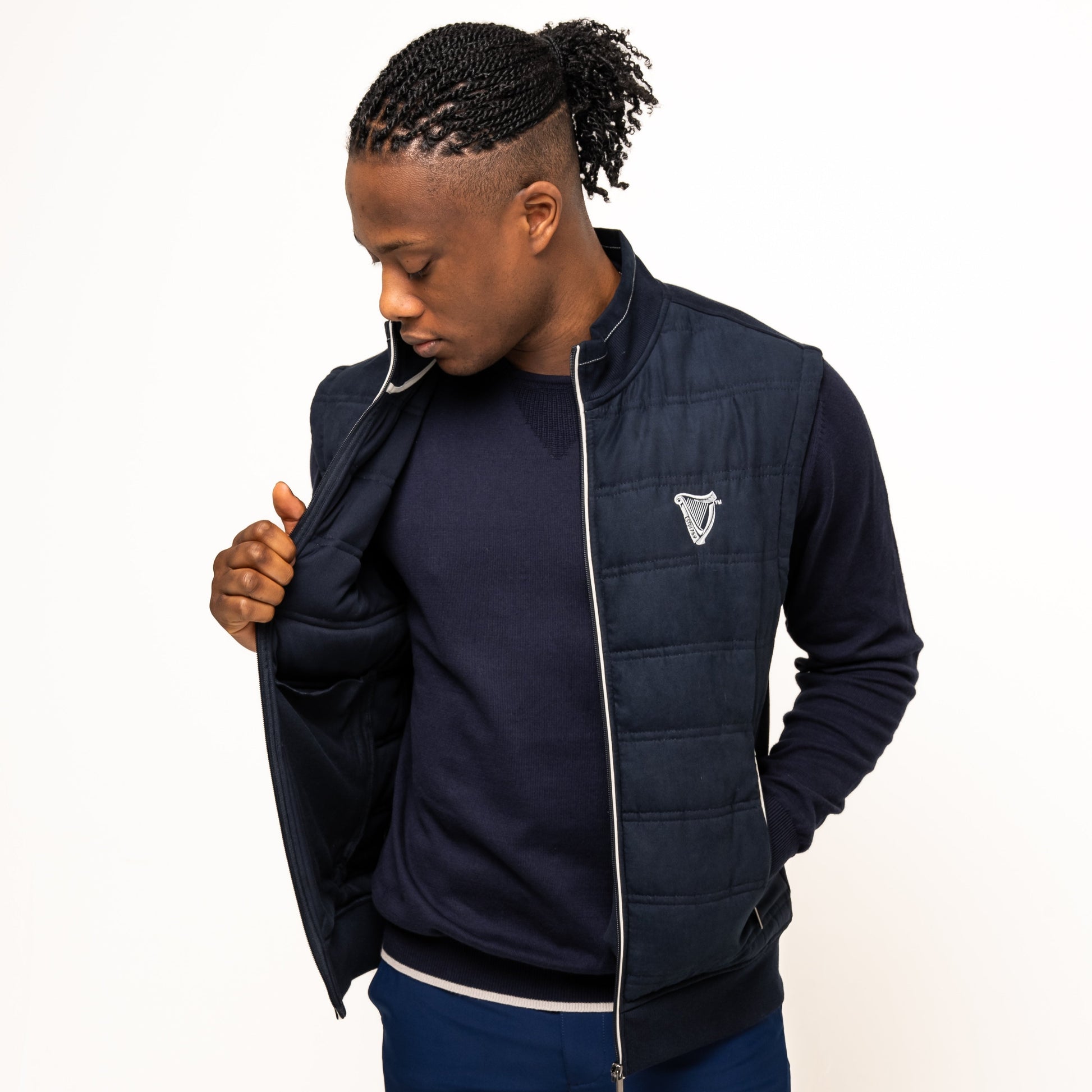 A man wearing the Guinness Performance Gilet from Guinness Webstore US, featuring quilted detailing, stands against a white background over a navy sweater and blue shorts, looking down and holding his vest open.