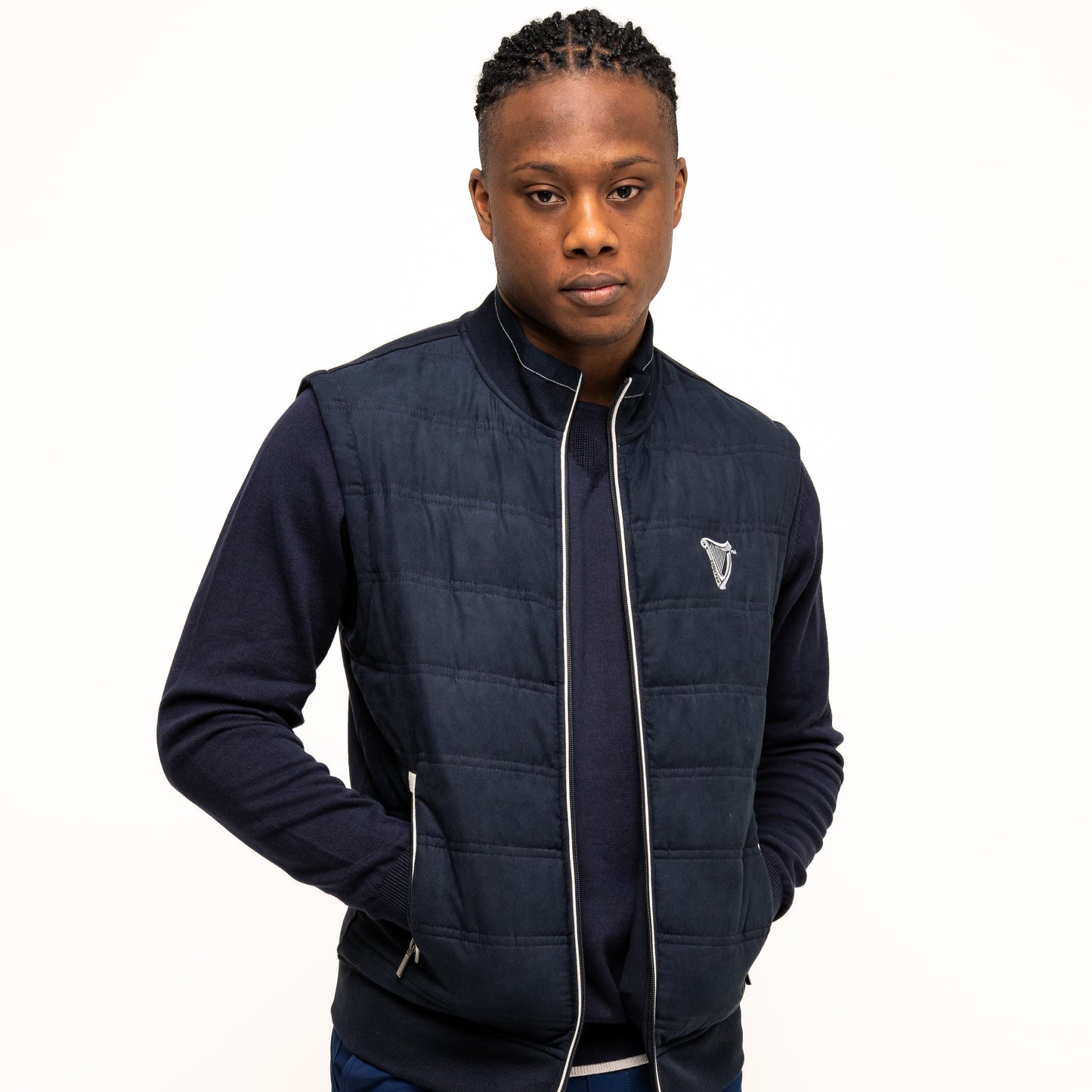 A man stands facing forward with his hands in his pockets, wearing a navy Guinness Performance Gilet from Guinness Webstore US over a long-sleeve shirt and navy shorts, set against a plain white background.