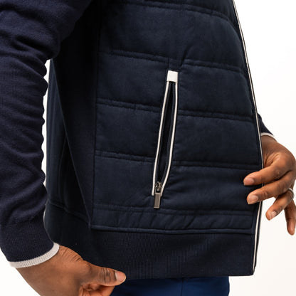 A person wears the navy Guinness Performance Gilet from Guinness Webstore US, featuring quilted detailing and blue shorts, with one hand near the gilet's zipper pocket and the other resting at their side.