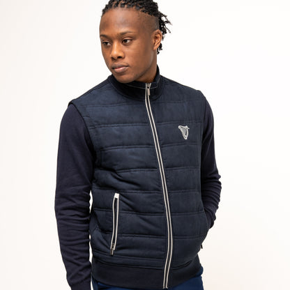 A person models the navy Guinness Performance Gilet by Guinness Webstore US, featuring quilted detailing, over a dark long-sleeve shirt and blue shorts against a plain white background.