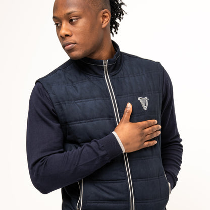 A person in the Guinness Performance Gilet from Guinness Webstore US, featuring quilted detailing, touches their chest with one hand while looking off to the side against a plain background.