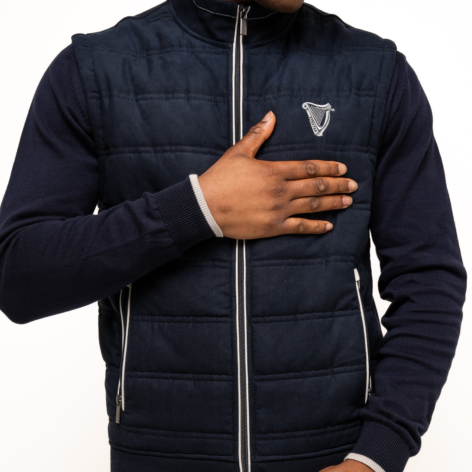 Wearing the Guinness Webstore US Performance Gilet with quilted details and logo zippered pockets, a person places one hand on their chest and the other on their jacket.
