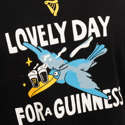 The Guinness Seb Curi Oversize Tee - Black from Guinness Webstore US features a cartoon blue bird flying with two pints of Guinness and the phrase "Lovely Day for a Guinness" on an oversized black tee.