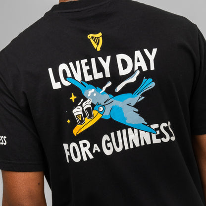 Someone wears the Guinness Seb Curi Oversize Tee - Black from Guinness Webstore US, featuring a blue bird with two pint glasses and the text "Lovely day for a Guinness.