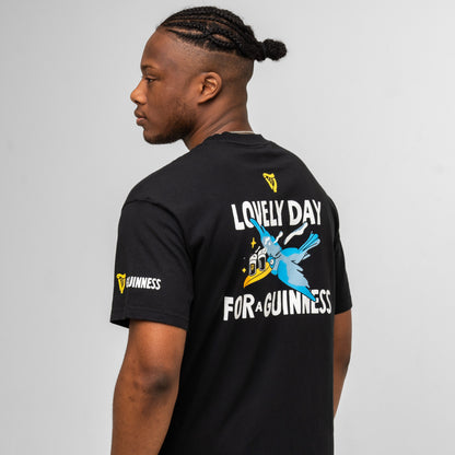 A man faces away, wearing the Guinness Seb Curi Oversize Tee - Black from Guinness Webstore US, featuring “Lovely Day for a Guinness” and a flying toucan graphic on the back, plus logos on the sleeve and upper back.